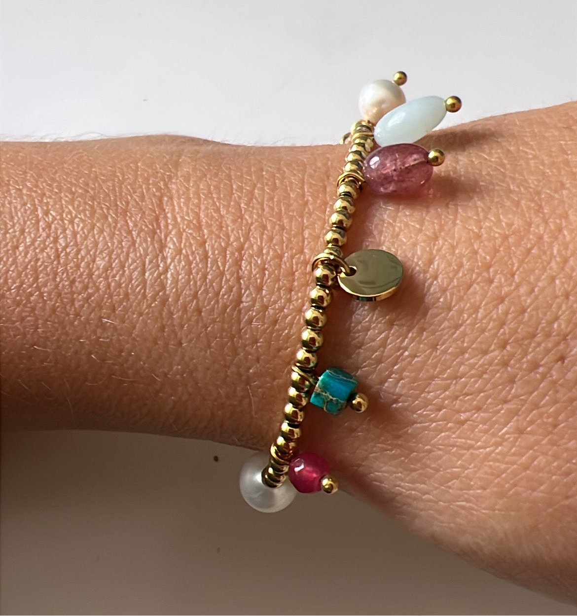 pulsera elástica waterproof o resistente al agua confeccionada en acero con baño de oro 18 kilates. Tiene abalorios de colores imitando piedra natural y perla cultivada. 
