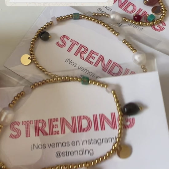 pulsera elástica waterproof o resistente al agua confeccionada en acero con baño de oro 18 kilates. Tiene abalorios de colores imitando piedra natural y perla cultivada. 