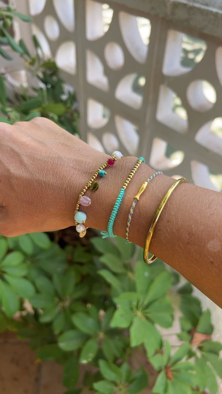 pulsera elástica waterproof o resistente al agua confeccionada en acero con baño de oro 18 kilates. Tiene abalorios de colores imitando piedra natural y perla cultivada. 